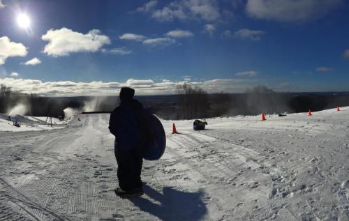 A person in winter gear stands on a snowy slope, with snowmaking machines and orange cones in the background under a bright sky.