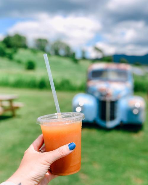 A hand holds a refreshing drink in a clear cup, with a vintage blue truck and green landscape in the background.
