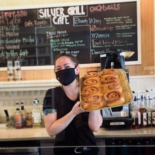 A masked barista holds a tray of cinnamon rolls in front of a café menu board.