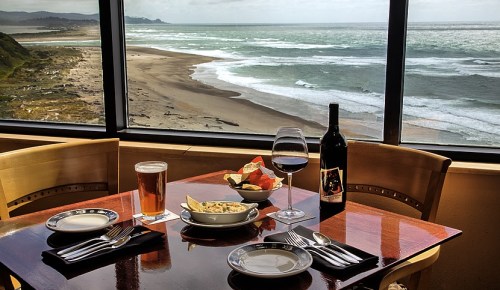 A dining table with a view of the ocean, featuring a glass of wine, a beer, and a plate of food.