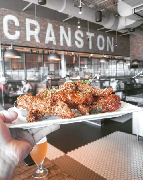 A hand holds a plate of crispy chicken wings in a restaurant with a "Cranston" sign in the background.
