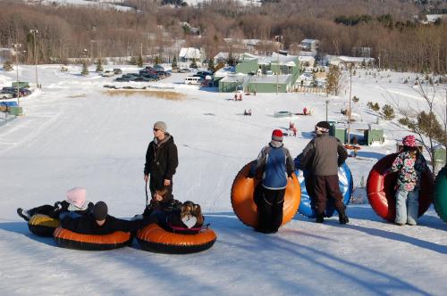 A snowy hill with people preparing to go tubing, surrounded by trees and buildings in the background.