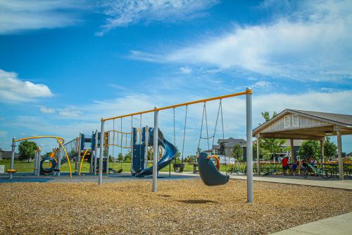A playground with swings, climbing structures, and a picnic shelter under a blue sky with scattered clouds.