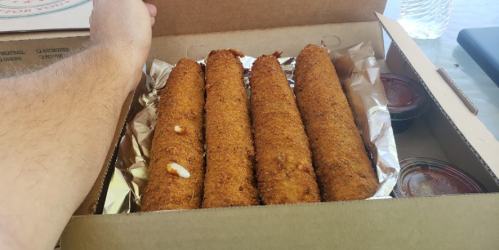 A hand reaches for a box containing four golden-brown, crispy rolls with two small containers of sauce.