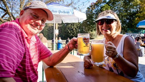 A man and woman toast with large beer mugs at an outdoor table, smiling in a sunny setting.