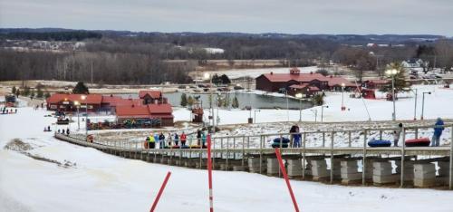 A snowy landscape with people tubing down a slope, surrounded by trees and buildings in the background.