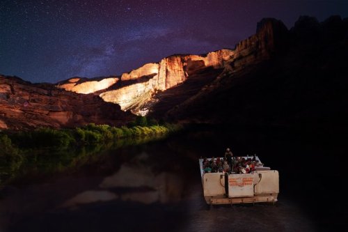 A boat with passengers floats on a dark river, surrounded by illuminated cliffs under a starry night sky.