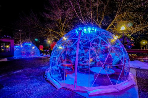 Colorful igloo-like structures illuminated at night, surrounded by trees and outdoor seating in a winter setting.
