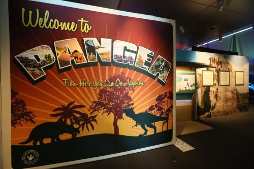 A colorful sign reading "Welcome to Pangea" with dinosaur silhouettes and a museum exhibit in the background.