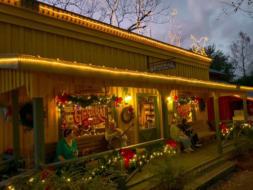 A festive general store adorned with lights and decorations, with people sitting on the porch at dusk.