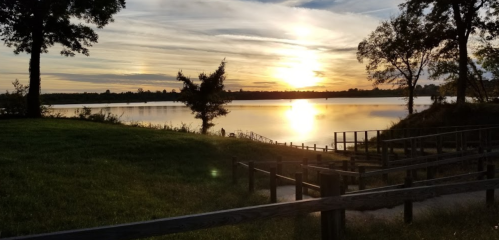 Sunset over a calm lake, with trees and a wooden walkway in the foreground, reflecting warm colors on the water.