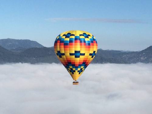A colorful hot air balloon floats above a sea of clouds with mountains in the background under a clear blue sky.