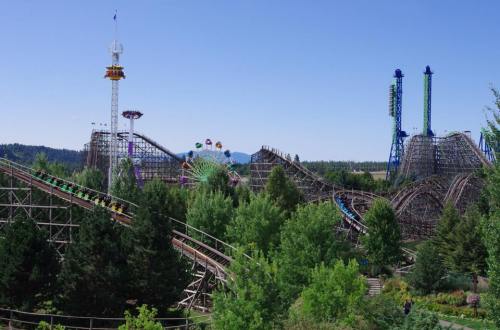 A vibrant amusement park scene featuring roller coasters, a Ferris wheel, and lush green trees under a clear blue sky.