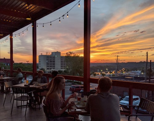 A sunset view from a restaurant patio, with diners enjoying drinks and a vibrant sky filled with orange and purple hues.