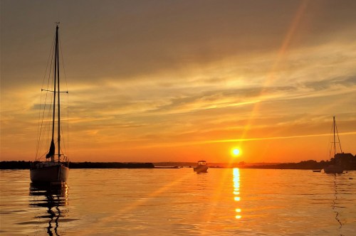 A serene sunset over calm waters, with sailboats silhouetted against the vibrant orange and yellow sky.