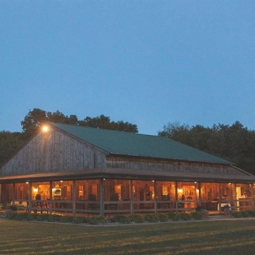 A rustic wooden cabin with a green roof, warmly lit at dusk, surrounded by trees and a grassy field.
