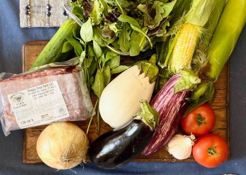 A wooden board with fresh vegetables: lettuce, corn, eggplants, tomatoes, onions, garlic, and a package of ground beef.