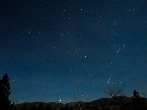 A starry night sky with scattered stars, a faint meteor streak, and silhouetted mountains in the background.