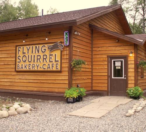 A wooden bakery-café with a sign reading "Flying Squirrel Bakery-Café" and an open sign, surrounded by gravel and plants.