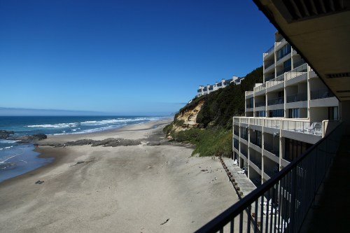 View of a sandy beach and ocean from a balcony, with cliffs and a hotel building in the foreground under a clear blue sky.