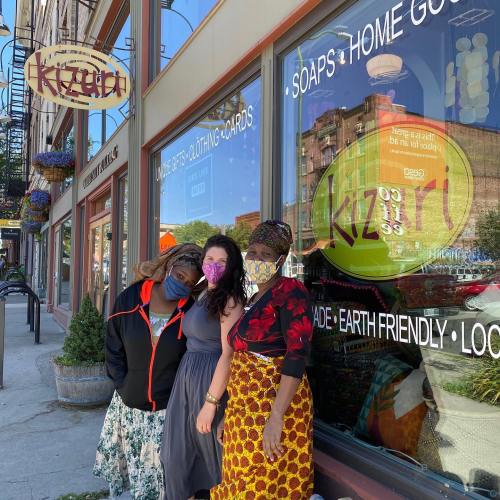 Four people pose outside a store called Kiziri, wearing masks and colorful clothing, with shop windows in the background.