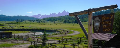 A scenic view of Buffalo Valley Ranch with mountains in the background, green fields, and a wooden welcome sign.