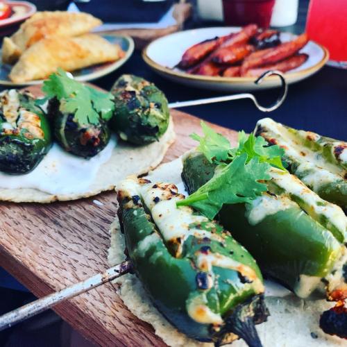 A wooden platter with grilled jalapeños stuffed with cheese, garnished with cilantro, alongside empanadas and sausage.