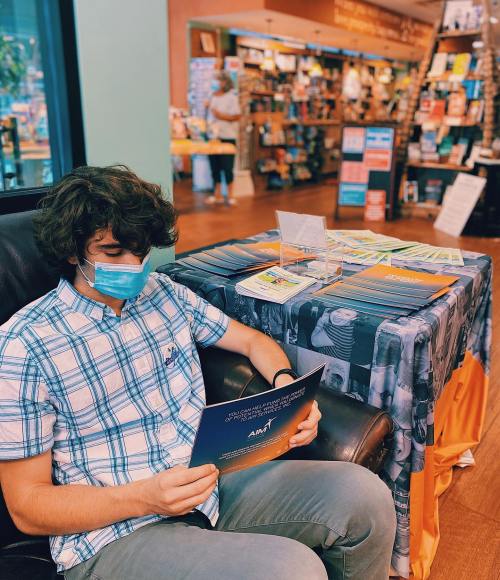 A young man in a mask sits in a bookstore, reading a brochure while surrounded by books and a display table.
