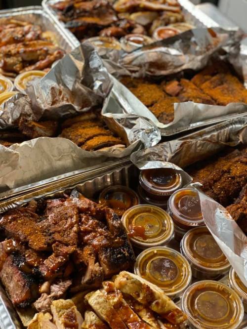 A variety of barbecued meats in foil trays, accompanied by small containers of sauce.