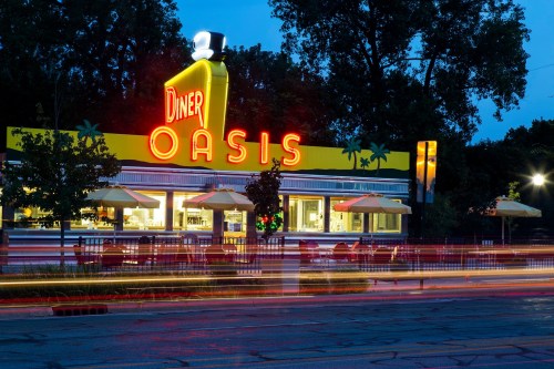 A brightly lit diner named "Oasis" at night, featuring neon signs and outdoor seating under umbrellas.