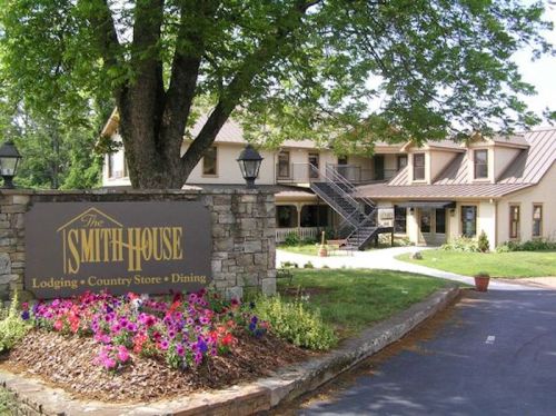 A charming country inn with a stone sign, surrounded by flowers and trees, featuring a two-story building and outdoor seating.