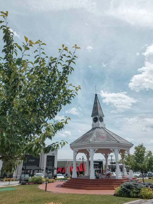 A gazebo with a clock tower surrounded by greenery and shops under a partly cloudy sky.