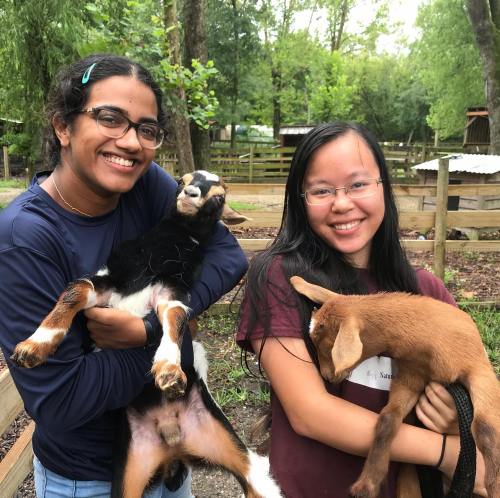Two smiling young women hold goats in a farm setting, surrounded by trees and wooden fencing.