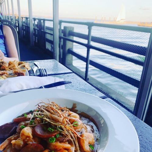 A plate of shrimp and sausage sits on a table with a waterfront view and a sailboat in the background.