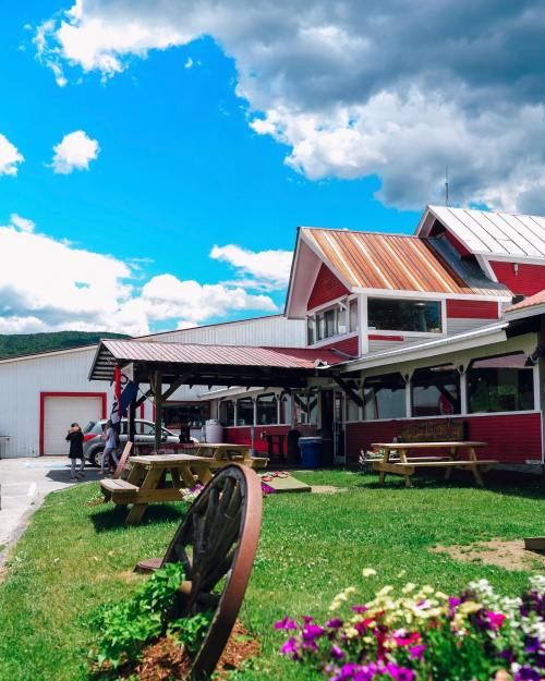 A red and white building with a metal roof, surrounded by green grass, picnic tables, and colorful flowers under a blue sky.