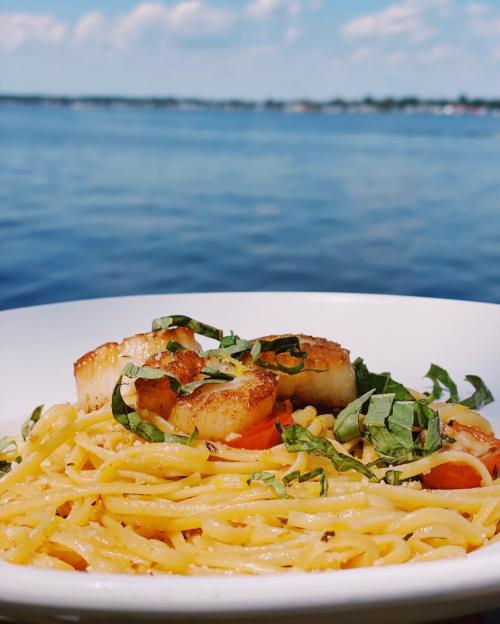 A plate of pasta with scallops and basil, set against a scenic waterfront view.