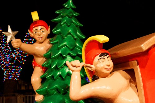 Two colorful elf statues playfully pose near a green Christmas tree, with festive lights in the background.