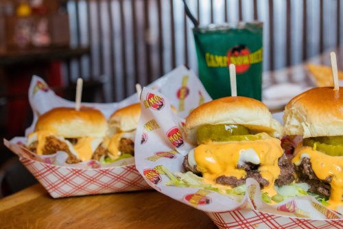Two sliders with cheese, pickles, and lettuce in a basket, alongside a colorful drink in the background.