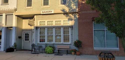 A quaint bakery storefront with an "OPEN" sign, outdoor seating, and potted plants on a sunny day.
