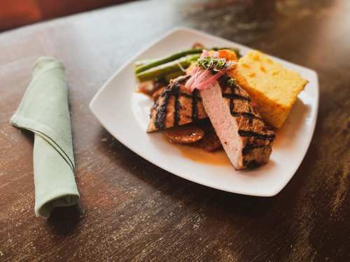 Grilled pork steak with asparagus, cornbread, and pickled onions on a white plate, accompanied by a green napkin.