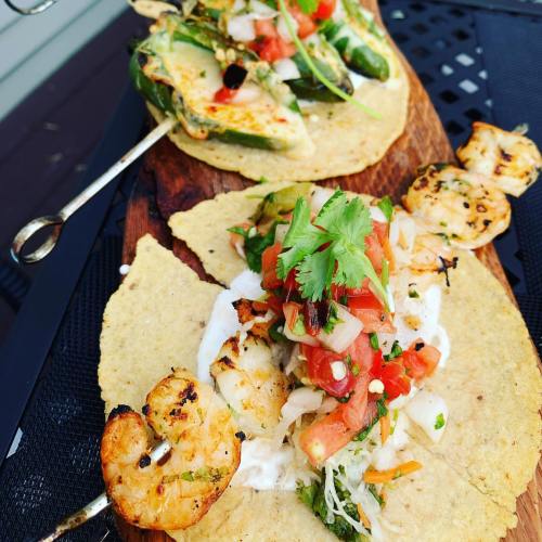 Two tacos on a wooden board, one topped with grilled shrimp, pico de gallo, and cilantro, the other with jalapeños and cheese.