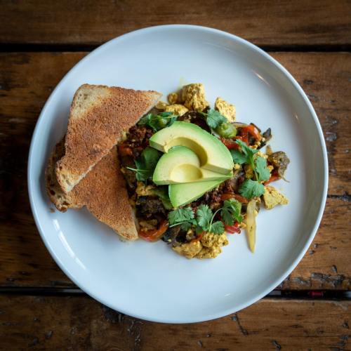 A plate of scrambled eggs with vegetables, topped with avocado slices, served with two slices of toasted bread.