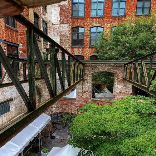 A view of a brick courtyard with green trees and a metal walkway connecting two buildings.