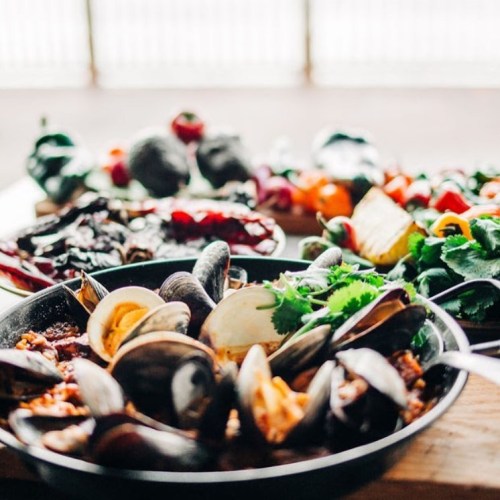 A close-up of a seafood dish with mussels, surrounded by colorful vegetables on a wooden table.