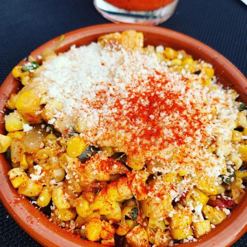 A clay dish filled with seasoned corn topped with cheese and paprika, set on a textured black surface.