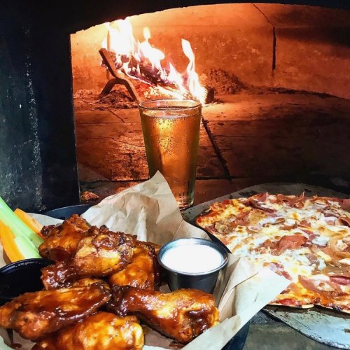 A plate of chicken wings with celery, a dipping sauce, and a pizza, with a glass of beer and a fire in the background.