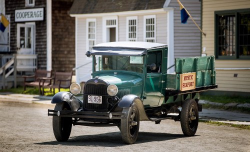 A vintage green truck parked on a dirt road in a historic seaport setting.