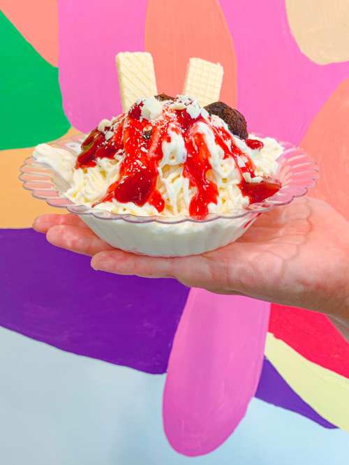 A hand holding a bowl of ice cream topped with whipped cream, chocolate cookies, and strawberry sauce against a colorful background.