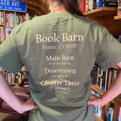 A person wearing a green t-shirt with "Book Barn" and location details printed on the back, standing in a library.