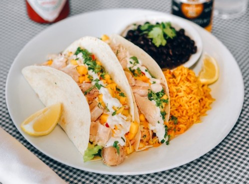 Three tacos filled with chicken, mango, and cilantro, served with yellow rice, black beans, and lemon wedges.